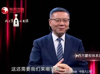 这是一张东方卫视节目的截图，画面中一位穿着蓝色西装、戴着眼镜的男性正在讲话。他打着蓝色条纹领带，佩戴着耳麦，双手交叠放在身前。背景为深色，左上角有东方卫视的标志和一个白酒品牌的广告。画面底部有文字"这还需要我们来嘲笑吗"，右侧有部分文字"西方霸权体系"。