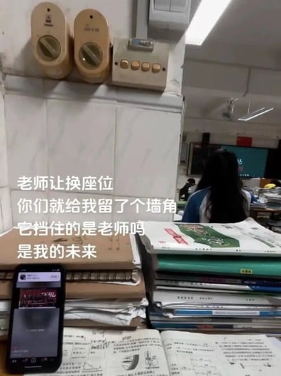 图片展示了一间教室的角落场景，前景是堆满课本和练习册的课桌，桌上还放着一部正在播放内容的手机。背景中可见其他学生和黑板，墙上有两个米色的挂式设备和一个控制开关。图片左侧叠加了一段文字：“老师让换座位 你们就给我留了个墙角 它挡住的是老师吗 是我的未来”，表达了学生被分到角落座位后的无奈与自嘲，通过夸张的比喻将物理空间的“挡住老师”延伸为对未来的担忧，充满校园生活的真实感和幽默感。