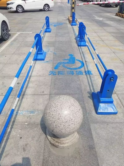 这张原始拍摄照片展示了一处户外公共区域的通道：灰色地砖地面上用蓝色油漆喷涂了轮椅标识和“无障碍通道”字样，明确标注此处为供行动不便人士使用的无障碍通道，但通道正前方设置了一个大型挡路石球，通道两侧也加装了蓝白相间的隔离护栏，整个通道被完全阻断，轮椅根本无法通行，形成了极具讽刺性的反差，暴露了不合理、形式主义的设施设计问题。