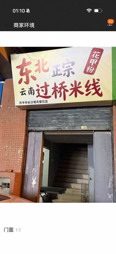 这是一张手机端商家展示页的截图，呈现了一家位于开平市的餐饮店门头，上方的店铺招牌出现了极具反差感的表述，将地域属性完全不相关的“东北”“云南”和“正宗”组合，宣传售卖过桥米线，同时还提供花甲粉，门店卷帘门半开，内部可见通往上层的楼梯，招牌的矛盾组合充满荒诞的幽默感，让人对该店米线的风味定位产生疑惑，具有很强的搞笑效果。