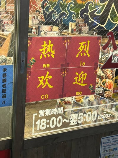 照片展示了一家商店的橱窗，橱窗上贴有红色背景、黄色文字的'热烈欢迎'标语，每个汉字下方配有疑似拼音的标注：'热(we)'、'烈(le)'、'欢(CO)'、'迎(me)'，形成有趣的组合。左侧有蓝色标牌写着'未经腺者歡迎!!'（疑似存在文字错误）。橱窗底部贴有白色营业时间告示，内容为日文：'営業時間 18:00~翌5:00 土日祝 12:00~'（营业时间：18:00至次日5:00，周末及节假日12:00开始）。背景中可见店内陈列的商品和带有波浪图案的装饰画，整体呈现出热闹的店铺氛围。