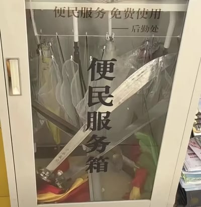 画面中是一个带玻璃门的金属柜，柜门上贴有“便民服务箱”五个大字，顶部印有“便民服务 免费使用 — 后勤处”的标语。柜子内部挂有多把雨伞，而在显眼位置却放置了一把金属砍刀，与常见的便民物品（如雨伞）形成强烈反差，营造出“砍刀是否属于便民服务”的幽默疑惑感。