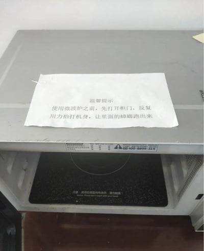 一张微波炉的照片，顶部放着一张写有幽默提示的白纸。提示内容为："温馨提示：使用微波炉之前，先打开柜门，反复用力拍打机身，让里面的蟑螂跑出来"。这张图片通过一本正经的"温馨提示"形式，用夸张的方式暗示微波炉可能成为蟑螂的藏身之处，制造出搞笑效果。