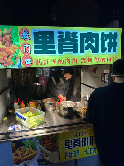 夜晚的街头美食摊位，上方亮着醒目的绿色招牌，主标题为“校门口 里脊肉饼”，左侧配有诱人的里脊肉饼图片，下方写着“满当当的肉肉 沉甸甸的回忆”，传递出满满的怀旧感。摊位前，一位戴着眼镜的摊主正在专注地制作食物，不锈钢操作台上摆放着装有生菜的透明盒子、多个金属盆和调料瓶，右侧有一位顾客正等待购买。摊位下方还有黄色招牌，印有“鼎顶香 里脊肉饼 饼酥肉香 一口过瘾”的宣传语，整体场景充满了市井生活气息和诱人的美食氛围。