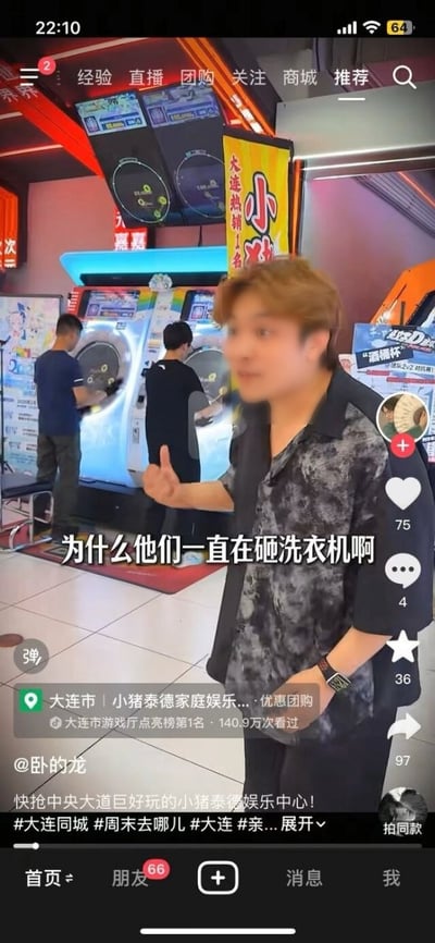 这是一张抖音短视频的竖屏截图，拍摄场景位于大连市的小猪泰德家庭娱乐游戏厅内。前景处一名黄发男子站在镜头前，面露疑惑的神情，一只手抬起做出发问的姿态，画面配文为“为什么他们一直在砸洗衣机啊”。背景中两台外观类似洗衣机的设备实际是游戏厅的音游设备，另有两名男子正在操作游玩该设备。界面上还显示了该地点是大连市游戏厅点亮榜第1名，累计有140.9万次看过，视频发布者为@卧的龙，附带大连同城、周末去哪儿等话题标签，右侧是抖音平台的点赞、评论、收藏、转发互动按钮，该视频属于同城探店推广内容，通过将音游设备误认成洗衣机的反差感制造笑点。