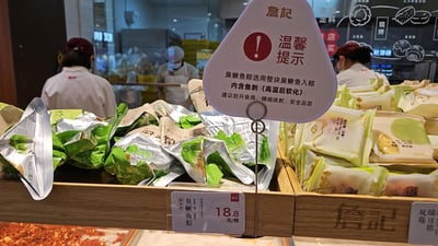 图片展示了詹記门店的食品陈列区，前景堆放着绿色和白色包装的粽子及点心，中央立着一个三角形温馨提示牌，牌面印有红色感叹号和“温馨提示”字样，详细说明臭鱖魚粽内含鱼刺需小心食用。背景中三位穿着白色工作服、戴帽子口罩的店员正在操作台后忙碌，整体呈现出中式点心店的日常销售场景，提示牌突出食品安全注意事项。