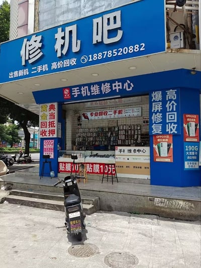 这是一张街头实拍的静态图片，展示了一家位于街边的手机维修类店铺。店铺主招牌为蓝色，顶部用醒目的白色大字标注店名“修机吧”，下方标注店铺业务包括出售新机、二手机、高价回收，同时留有联系电话。店铺入口处标有“手机维修中心”字样，店内外张贴了多块宣传标识，覆盖的业务还有爆屏修复、二手手机回收、黄金回收、典当抵押、10元手机贴膜，同时还有OPPO手机促销、手机流量卡办理、宽带报装等相关业务宣传。店铺门前的台阶上摆放了数把高脚凳，台阶下方的人行道上停放着一辆电动自行车，背景可见街道旁的树木和其他街景元素。该店铺名称“修机吧”存在谐音歧义，是这张图的趣味核心。