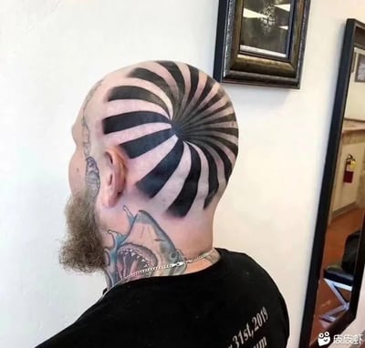 这是一张实拍的静态图，画面展示了一位留有浅棕色胡须的光头男性的侧后方，他的头顶纹有黑白色交替的放射状螺旋条纹纹身，该纹身会让人产生头部向内凹陷形成空洞的视觉错觉；他的侧脸和颈部也有多处纹身，其中颈部是鲨鱼张嘴的图案，他身穿黑色上衣，颈部佩戴银色链条项链，背景是室内的白色墙面，墙上挂有装饰画和镜子，图片右下角带有皮皮虾平台的水印。