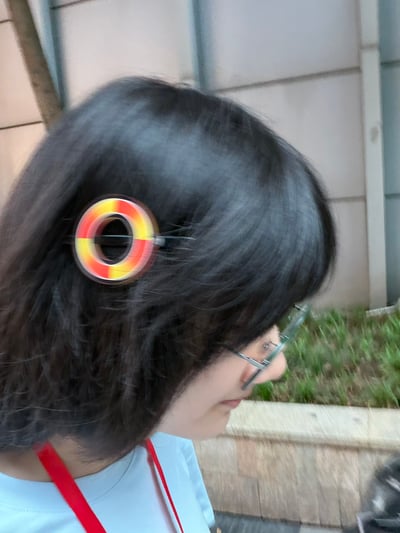 这是一张真人实拍的静态照片，画面主体是一名留黑色短发、佩戴金属框眼镜的女性的侧脸，她的头发上别着一个设计成视频加载时红黄渐变环形缓冲图标的发夹，视觉效果十分有梗，仿佛人物正在加载中。人物身穿浅色上衣，脖子上挂着红色挂绳，背景是户外的浅灰色建筑墙面、绿化带和路边石台，整体氛围轻松有趣，展现了创意小物件的趣味效果。