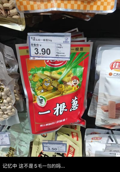 这是一张超市货架的实拍图，主体是光头娃品牌的「一根葱」怀旧零食，包装以红橙为主色调，印有卡通葱形象、新鲜葱和零食的图案，标注「非油炸」「原汁原味的儿时味道」。上方的盒马超市价签显示该56g装零食零售价为3.90元。图片底部配文「记忆中 这不是5毛一包的吗...」，直白表达出对童年零食价格大幅上涨的感慨，画面周围还可见花生、佳宝品牌等其他零食产品。