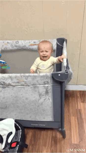 这是一段动态GIF，画面中一个小宝宝站在带有图案的灰色游戏围栏内，一只手紧紧抓着围栏的金属栏杆，张着嘴巴崩溃大哭，面部满是委屈的神情，场景位于室内铺有木地板的房间，背景是浅色的墙面，真实记录了宝宝的情绪瞬间。