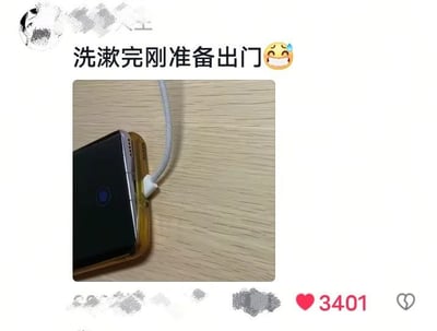 图片上方有文字“洗漱完刚准备出门😅”，下方是一张实物照片。照片中展示了一部黑色手机（带有金色边框），屏幕朝下放置在浅色木质桌面上，手机底部连接着一根白色的充电线，充电线另一端延伸出画面。手机侧面有一个蓝色的指示灯亮起，显示正在充电状态。整体场景呈现出一种日常生活中常见的小尴尬：当人们洗漱完毕、准备出门时，却发现手机还在充电，无法立即带走，配合文字中的流汗表情，生动传达了那种既无奈又哭笑不得的情绪。