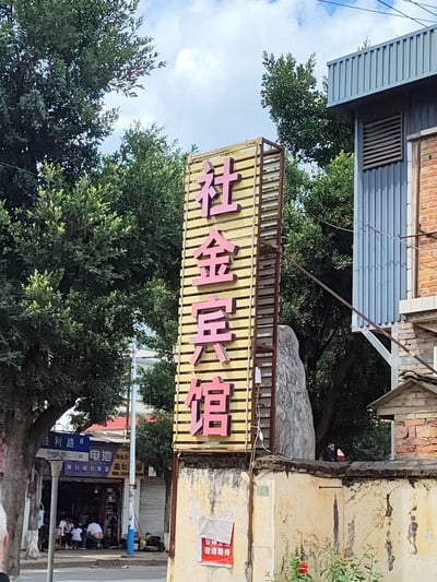 这是一张街头实拍的竖版照片，画面主体是黄底粉字的竖版“社金宾馆”招牌，招牌安装在一处老旧斑驳的墙面上，墙面还贴有红底白字的“暂停营业 敬请期待”告示。背景中可见茂盛的绿树，街边有一家标注“胜利路西”的电池店铺，店内有几位顾客就座，整体呈现出充满生活气息的城镇日常街景。