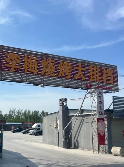 这是一张户外场景的实拍图，画面上方是一块大型的红底黄字招牌，醒目地写着“李梅烧烤大排档”，招牌右侧还有红色的联系电话字样。招牌下方右侧立着一块竖牌，标注“大车停车场”，竖牌下方搭配有红色的装饰图案。画面里能看到开阔的水泥停车场地，场地内停放着几辆轿车和一辆房车，旁边是灰色的简易建筑，背景是晴朗的蓝天和远处的树木，整体呈现出一个兼具餐饮服务与大型车辆停车功能的场地，看起来主要面向过路车辆尤其是长途大车司机，提供餐饮和停车的配套服务。