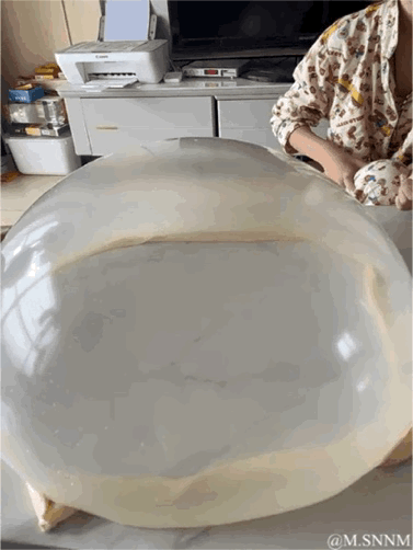 这是一段动态GIF图，画面中一位穿着花睡衣的人在室内展示自己揉制的面团，这张面团被擀得薄近乎透明，面积宽大，能清晰透过面团看到后方的柜子、打印机等家具，直观展现了高超的面食制作手艺，画面充满生活感，让人惊叹于这份精湛的厨艺。