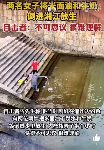这是一段社会事件的视频截图，画面呈现了湘江边的台阶区域，两名女子正在进行特殊的放生行为：其中一名身着深色外套的女子正将手中的物品倒入浑浊的湘江水中，另一名穿红色上衣的女子站在上方台阶，身旁摆放着米面油、矿泉水等准备用于放生的物资。图中通过多处文字对事件进行说明，顶部文字点明核心事件“两名女子将米面油和牛奶倒进湘江放生”，并标注目击者的态度“目击者：不可思议 很难理解”；底部文字则是目击者马先生的口述内容，他表示自己当时正在江边钓鱼，目睹两位阿姨花费约半小时将米面油、矿泉水、牛奶等物品倒入江中放生，他对这种行为感到无法理解。画面右侧带有“果然”的媒体水印，表明这是正规媒体对该事件的记录。