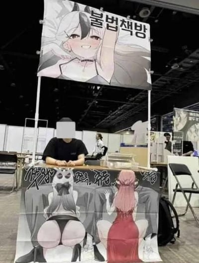 这是一张展会现场的实拍图，摊主利用视觉错位打造搞笑效果：桌子前方的布帘印有两名二次元女性的下半身，坐在桌后的人仅露出上半身，从正面看仿佛这个人的下半身就是布帘上的二次元臀部，形成趣味视觉错觉。摊位上方的横幅印有一名微笑抬手的二次元白发女性，横幅上的韩文“볼버채방”是谐音“불법채방”（非法聊天室）的玩笑梗，将“불법”（非法）替换为谐音的“볼”（此处指代臀部），玩起了“摸屁股房”的低俗趣味谐音，整体设计用来吸引展会游客的注意。