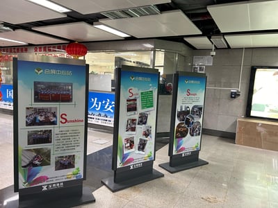 这是在深圳地铁会展中心站拍摄的线下宣传场景，站内设置了三块立式图文展示牌，分别以「Sunshine（阳光）」「Smile（微笑）」「Strength（力量）」为核心主题，展示了车站工作人员的团队活动、日常工作、服务乘客等场景照片，并配有详细文字介绍，传递阳光向上的团队文化与贴心服务的精神。背景可见车站的办公区域、红色装饰灯笼、墙面标语以及深圳地铁的官方标识，整体氛围正式且充满正能量，是地铁车站内的企业文化宣传场景。