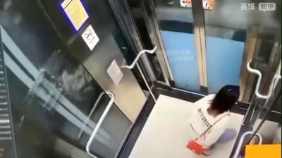 这是一张玻璃电梯内的监控视角截图，画面中一位背着红色挎包、穿着带有部分字母“...ORIST”白色上衣的女生蹲在电梯地面，身体背对镜头。电梯的玻璃壁上反射出一个模糊的黑影，其轮廓看起来像有着夸张面部的“怪物”，实际上这是由女生自身或电梯内物品反光形成的视觉错觉。女生的蹲坐姿态推测是被这个突如其来的“黑影怪物”吓到，整体场景是生活中一场乌龙又有点惊悚的巧合。