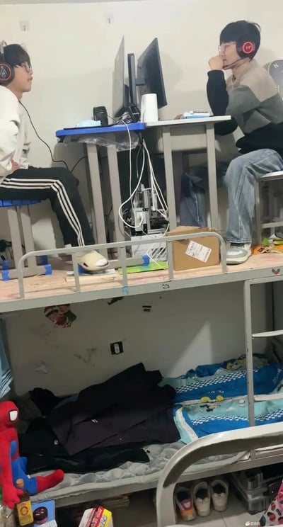 图片展示了一个学生宿舍，原本的上下铺床，上铺被改造成一张共享电脑桌，两位同学面对面坐着打游戏或使用电脑。桌上有耳机、显示器等电子设备，下铺仍为床铺，堆放着衣物和小物品，还有一个蜘蛛侠毛绒玩偶，空间紧凑但富有生活气息。整个画面传递出宿舍DIY创新精神与典型宅文化氛围，非常有真实感和幽默感。

画面没有明显谐音梗，但场景反转与空间创新本身具有一定幽默与反差效果。
