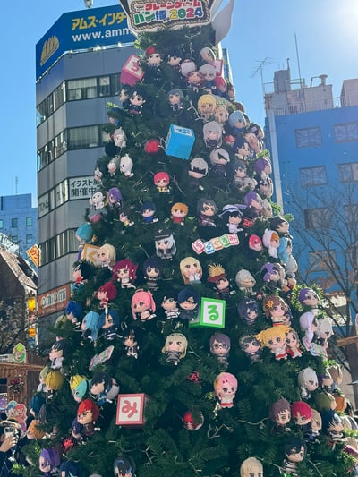 这是在日本东京秋叶原街头拍摄的特色圣诞树，整棵树上密集挂满了来自不同二次元作品的Q版角色毛绒挂件，搭配带有日文假名的方块装饰，充满创意。背景是秋叶原标志性的动漫主题店铺，顶部设有「クレーンゲームパン博2024」（抓娃娃主题博览会2024）的标识，是年末展会的圣诞主题布置，浓厚的二次元氛围吸引大量动漫爱好者前来打卡。