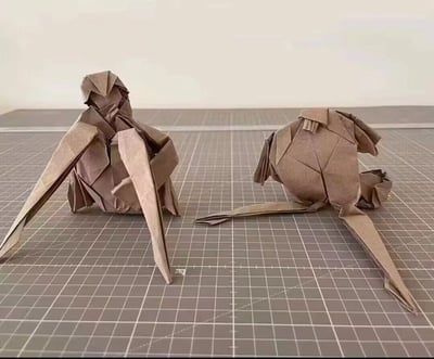 图片展示了两个用棕色纸张折叠而成的 origami 动物摆件，放置在带有白色网格线的灰色切割垫上。左侧的折纸造型较为复杂，具有明显的翅膀和头部结构，右侧的则相对圆润，两者均通过精细的折叠工艺呈现出立体感。背景为简洁的浅色，突出展示折纸作品的细节和纹理。