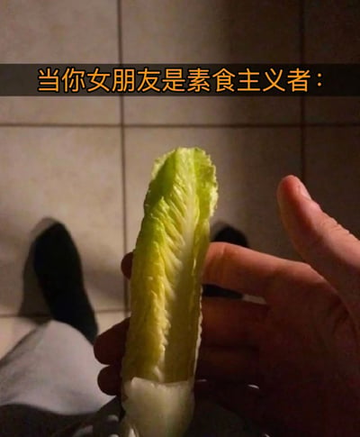 一张生活场景照片，展示了一只手拿着一片生菜叶，背景是瓷砖地面和一双穿着黑色袜子的脚。图片上方有黄色文字："当你女朋友是素食主义者："，通过展示直接拿着生菜叶的场景，幽默地调侃了与素食主义者伴侣相处时可能遇到的饮食情况。
