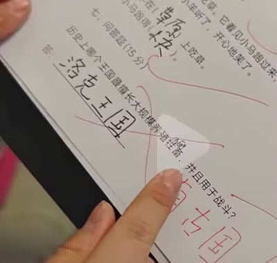这是一张实拍的小学生作业纸的照片，一只手拿着作业，手指指向作业的问答题区域。问答题题目为“历史上哪个王国最擅长大规模养殖牲畜，并且用于战斗？”，学生填写的答案是“洛克王国”，该答案被老师用红笔划叉批改。作业上方的其他填空也有答错被打叉的内容，这个答案利用谐音玩梗，“洛克”谐音“骡克”，对应题目中养殖骡马这类牲畜用于战斗的描述，是充满趣味的错误答题内容，十分搞笑。
