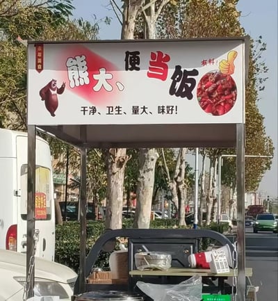 这是一张街边移动餐车的实拍静态图，画面主体是餐车上方的白底宣传招牌：左侧印有国产动画《熊出没》的角色熊大的卡通形象，招牌主文字为「熊大、便当饭」，右侧标注「有料！」字样和点赞手势图案，搭配红色餐品的实拍圆形展示图，招牌下方印有宣传语「干净、卫生、量大、味好！」，左上角还有竖排的「全年美食」红色标识。该招牌的文字排版存在断句歧义，很容易被误读为谐音梗「熊大便 当饭」，笑点十足。背景是城市户外街景，可见成排的行道树、路边停放和行驶的车辆，餐车台面上摆放着打包餐盒、扩音喇叭、收款二维码牌等经营用具。