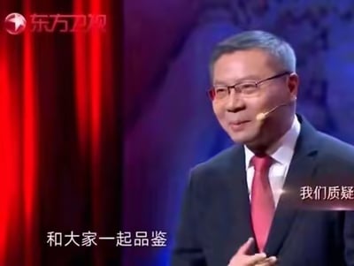 这是东方卫视某节目的视频截图，画面中的人物是学者张维为，他身着深色西装搭配红色领带，佩戴着演讲耳麦，面带微笑，神态亲和，双手做出手势，似乎正在进行观点分享与演讲。画面配有字幕“和大家一起品鉴”“我们质疑”，背景是红紫色的舞台幕布，整体呈现出正式的演讲场景氛围。