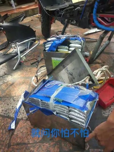 图片拍摄于室外的街边场地，地面上摆放着两个不锈钢箱体，里面塞满了用铝箔和蓝色包装材料包裹的自制电池组，电池组的线路裸露在外，看起来是为电动车改装的大容量电池。旁边是一辆被拆解了部分外壳的电动车，右下角有一只手正指向电池组。图片底部配有蓝色文字“就问你怕不怕”，用来调侃这种私自改装大容量电池的行为存在极大的安全隐患，这类没有正规防护和温控设计的自制电池组，很容易在使用过程中出现起火、爆炸等危险事故，图中场景大概率是在电动车维修或改装的街边店铺，用来警示人们不要进行这类违规且危险的操作。