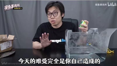 这是B站UP主“摸鱼事务所”的视频截图，画面中UP主坐在黑色桌前，面前摆放着透明容器和纸箱，一只手抬起，另一只手拿着小物件，表情无奈，配文“今天的难受完全是你自己造成的”，适合用来吐槽那些自作自受的行为，图中没有双关、谐音梗或暗示内容。