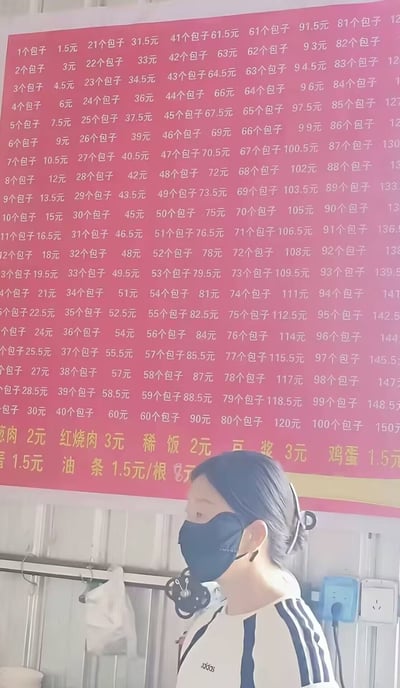这张实拍图拍摄于餐饮店铺内，画面上半部分是红色背景的餐品价目表，最反常的是商家没有直接标注包子的单价，反而将购买1到100个包子对应的全部总价逐一罗列，计算可知包子单价为1.5元/个，价目表下方还标注了卤肉、红烧肉、稀饭、豆浆、鸡蛋、油条等其他餐品的单价；画面下半部分是一位佩戴黑色口罩、身穿黑白条纹上衣的女士，站在价目表下方侧身看向左侧。该价目表因过于繁琐多余的标注方式成为笑点。
