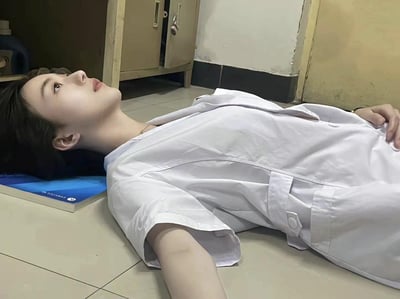 图片中是一位身穿白大褂的人，躺在地板上，姿势显得极为疲惫和无助，旁边是类似实验室或办公室的环境。此场景非常符合医学、科研等高压行业员工的常见“累瘫”状态，有一种“工作压垮人”的含义。地上铺着一本书当枕头，细节突显了身心俱疲又不得已临时休息的窘迫。整体透露出一种“崩溃又自嘲”的氛围，常被用作形容加班、熬夜、疲劳到极限的状态。