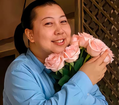 图片中是一位穿着蓝色衬衫的人物，面带灿烂微笑，双手捧着一束粉色玫瑰花。背景中可以看到带有几何图案的金属装饰结构，整体画面呈现出温暖愉悦的氛围。人物的笑容自然亲切，玫瑰花束新鲜饱满，粉色的花朵与蓝色的衬衫形成了和谐的色彩对比。