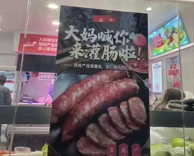 图片展示了一个食品店的广告海报，主要宣传猪肉灌肠产品。海报中央有大幅香肠图片，上方醒目文字写着“大妈喊你来灌肠啦!”，下方小字标注“现挑严选现灌装，放心猪肉放心肠”。背景中可以看到店铺内部环境，有其他食品广告和标语，包括“现挑严选放心猪肉”等宣传语。整体设计以红色和黑色为主色调，突出食品的新鲜和品质。