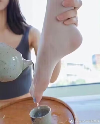 图片中一名女子正用脚拿着茶壶往杯子里倒茶，展现了一种非常规且令人惊讶的倒茶方式。女子留着长发，穿着深色背心，背景中可以看到窗外的城市建筑。这一画面通过使用脚部而非手部进行精细动作，创造出一种幽默且令人迷惑的视觉效果。