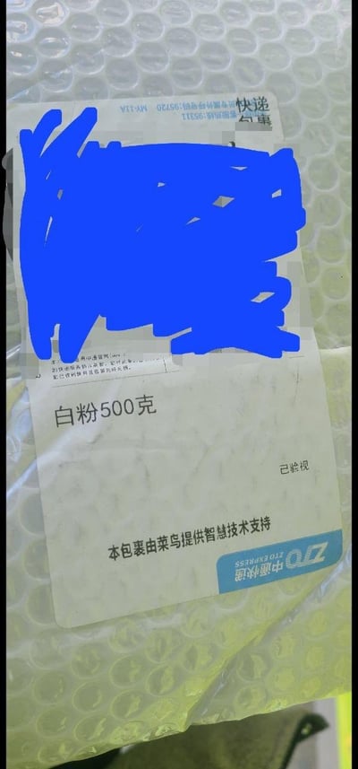 一张快递包裹的照片，包裹表面贴有物流标签。标签上有明显的蓝色涂鸦覆盖了部分信息，可见文字包括'白粉500克'、'已签收'以及'本包裹由菜乌提供智慧技术支持'。背景为气泡膜包装材料。

图片中'白粉'一词存在双关含义，字面意思是白色粉末，但在中文语境中也可能被理解为毒品的隐语，形成一种幽默或误导性的效果。
