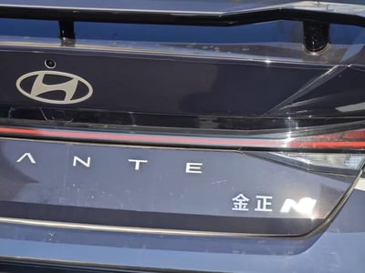图片展示了一辆现代汽车的尾部特写，中央位置可见现代汽车标志性的银色椭圆'H' logo，下方有大写字母'ANTE'字样。右侧区域则出现中文'金正'与字母'M'组合的标识，整体画面聚焦于车尾设计细节，背景为车身金属质感表面，光线反射清晰。