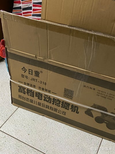 这是一张室内原始拍摄的照片，展示了「今日童」品牌型号为JRT-318的高档电动挖掘机玩具的外包装纸箱。纸箱为棕色硬纸板材质，顶部已被打开，能看到内部带有红、橙、黑、白相间条纹图案的包装布料。纸箱侧面清晰印有品牌标识「今日童®」、型号「JRT-318」、宣传语「金牌品质 恋童铸就」、生产企业「邢台恋童儿童玩具有限公司」，同时标注了执行标准：GB 6675.1-2014、GB 6675.2-2014、GB 19865 2005，还有一个标注「挖机安装视频」的二维码，以及挖掘机玩具的轮廓图案。地面是带有浅色斑点的瓷砖，纸箱左侧边缘能看到部分红色塑料袋和蓝色容器的边角。需要说明的是，宣传语中的「恋童」为生产企业名称的组成部分，属于品牌相关命名，无不良引申含义，不存在双关或谐音梗。