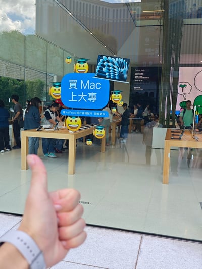 照片展示了一家苹果零售店的玻璃外墙，上面贴着蓝色促销海报，写着'买Mac上大学'并配有戴学士帽的笑脸表情，旁边还有MacBook的图案。海报下方小字写着'选AirPods或合资格配件，即省更多'。店内可以看到顾客正在体验产品，前景有一只手竖起大拇指点赞。