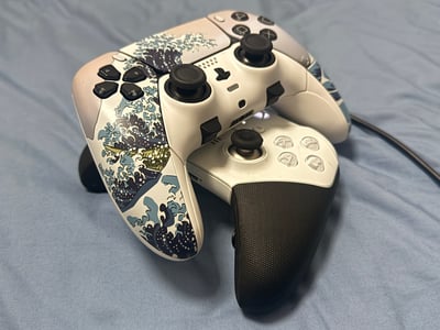 图片展示了两款高端游戏手柄的叠放对比，上方为PlayStation DualSense Edge手柄，机身印有《神奈川冲浪里》风格的蓝白海浪浮世绘图案，黑色摇杆与功能键点缀其间；下方为Xbox Elite手柄，采用白色主体搭配黑色防滑握把，按键布局清晰。两款手柄均放置于浅蓝色布料背景上，线缆自然垂落，整体构图聚焦于手柄的外观设计细节，疑似用于后续的性能评测对比内容。