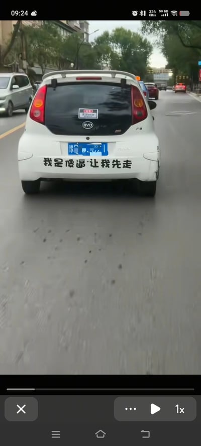 这是一张城市街道上的视频截图，画面主体为一辆白色比亚迪汽车的尾部。车辆行驶在车流量适中的道路上，背景可见树木和其他车辆。最引人注目的是车尾保险杠上贴着的黑色文字贴纸，内容为“我是傻逼让我先走”，采用自嘲式幽默的方式表达，可能是车主为了缓解驾驶压力或博人一笑的创意贴纸。