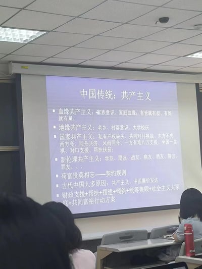 一张教室场景的照片，前方投影仪屏幕上显示着标题为“中国传统：共产主义”的PPT内容，列举了“血缘共产主义”“地缘共产主义”“国家共产主义”等多个分类及相关描述，台下有学生背影。