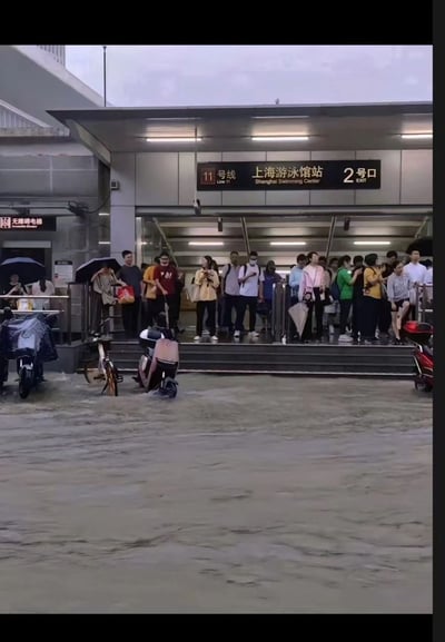上海地铁11号线上海游泳馆站2号口，雨天有大量人群聚集在出站口。许多人打着雨伞，地面湿滑，入口处停放着几辆电动车和自行车。人们或站或坐，似乎在等待或避雨，展现了城市通勤者在雨天的日常场景。