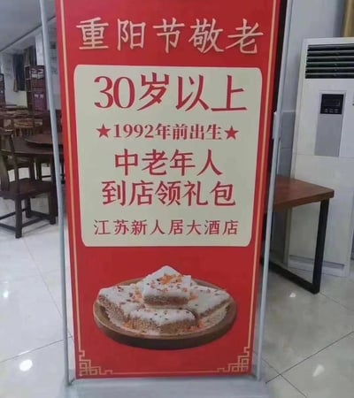 这是一张实拍的江苏新人居大酒店重阳节敬老活动宣传展架照片，展架为红色竖版易拉宝样式，放置在餐饮门店内部，背景可见店内的木质桌椅和立式空调。展架顶部标注“重阳节敬老”，中间区域说明活动规则：将30岁以上（1992年前出生）的人群定义为中老年人，符合条件者可到店领取礼包，展架底部还配有一份撒有配料的重阳糕类点心的图片。该宣传内容因将30岁人群归类为中老年人，与大众普遍认知的中老年人年龄标准存在极大偏差，引发网友调侃传播。