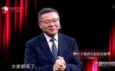 图片为电视节目《这就是中国》的截图，画面中一位身着深蓝色西装、佩戴条纹领带的男性正站在红色幕布前，面带微笑，双手交叠于身前。屏幕左下角有文字“大家都笑了”，右侧有“一个演讲引起的反响”字样，显示演讲内容引发了现场笑声。整体场景正式且带有轻松氛围，人物表情温和。