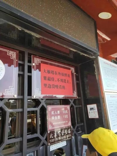 这张图片拍摄的是西安大雁塔相关售票点的窗口场景，窗口处悬挂着多块带中式边框的提示牌：最上方的红色提示牌写着一句十分实诚的提示“大雁塔在外拍照效果最佳，不用进去，里边没啥看的”，打破了常规景点引导游客购票入园的思路；下方的票价牌标注了“成人票 30元，学生票 15元”，并补充“凭票当次有效并直接入寺”，还有一块黑底白字的牌子用中英文标注“此票不含登塔 TICKET FOR THE PAGODA TOWER”。窗口右侧还贴有支付宝支付标识和其他说明告示，画面右下角能看到一个戴着黄色帽子的人的部分头部，整体场景充满接地气的趣味感。