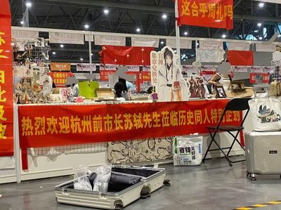这是国内某同人漫展的历史同人主题摊位现场，摊位前方悬挂醒目的红色横幅，用现代调侃的语气写着“热烈欢迎杭州前市长苏轼先生莅临历史同人特别企划”，玩了时空错位的搞笑梗——苏轼曾在杭州任知州，被爱好者用“前市长”的现代职务称呼。摊位内设有苏轼的古风同人立牌，标注“官摊 集章兑换处”，摊位上方还挂着红色横幅“这合乎周礼吗”，摊位上摆放着各类历史同人周边，周围还有其他漫展摊位，整体氛围活泼有趣，是爱好者们的趣味整活现场。