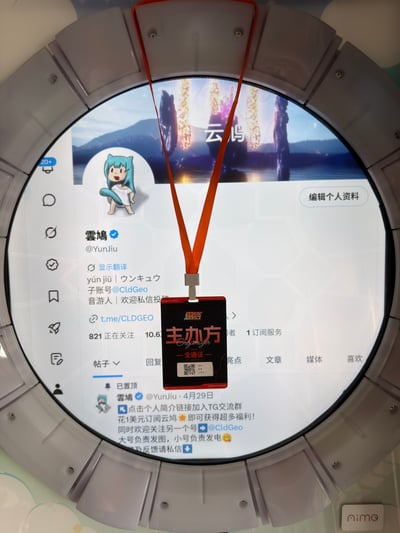 这是一张原始实拍照片，画面核心是一块灰色拼接边框的圆形显示屏，屏幕上展示了X（原推特）平台用户“雲鳩（@YunJiu）”的个人主页，主页头像是蓝发卡通形象，简介标注了拼音、日文译名、子账号@CldGeo、身份为音游人，还附有Telegram交流群链接，主页显示其关注数821、粉丝数约10.6万，置顶帖引导用户加入交流群、付费订阅获取福利，同时说明大号负责发图、小号负责发电。屏幕前方悬挂着一条橙色挂绳的黑色工作牌，工作牌上标注有“主办方 全通证”字样及二维码，遮挡了主页的部分内容，屏幕右下角可见“nimo”标识。